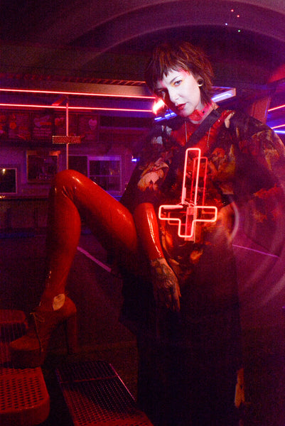 Katrina models a neon uv pink choker by Apatico and Anhedonie, a floral dress by Samantha Rei, and a red neon cross light under blue and red neon lights at night.