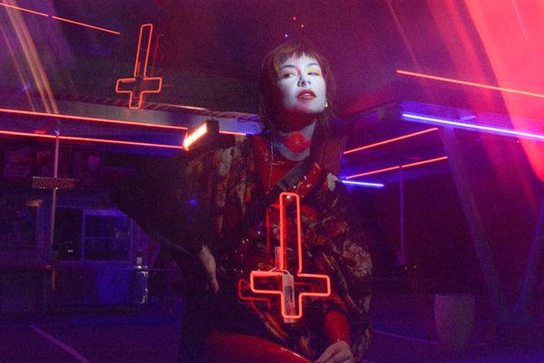 Katrina models a neon uv pink choker by Apatico and Anhedonie, a floral dress by Samantha Rei, and a red neon cross light under blue and red neon lights at night.