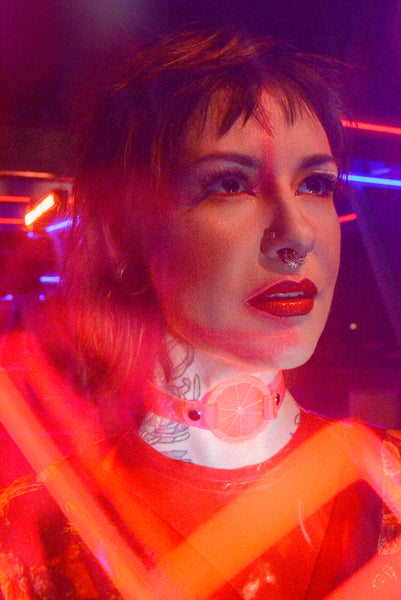 Katrina models a neon uv pink choker by Apatico and Anhedonie, a floral dress by Samantha Rei, and a red neon cross light under blue and red neon lights at night.