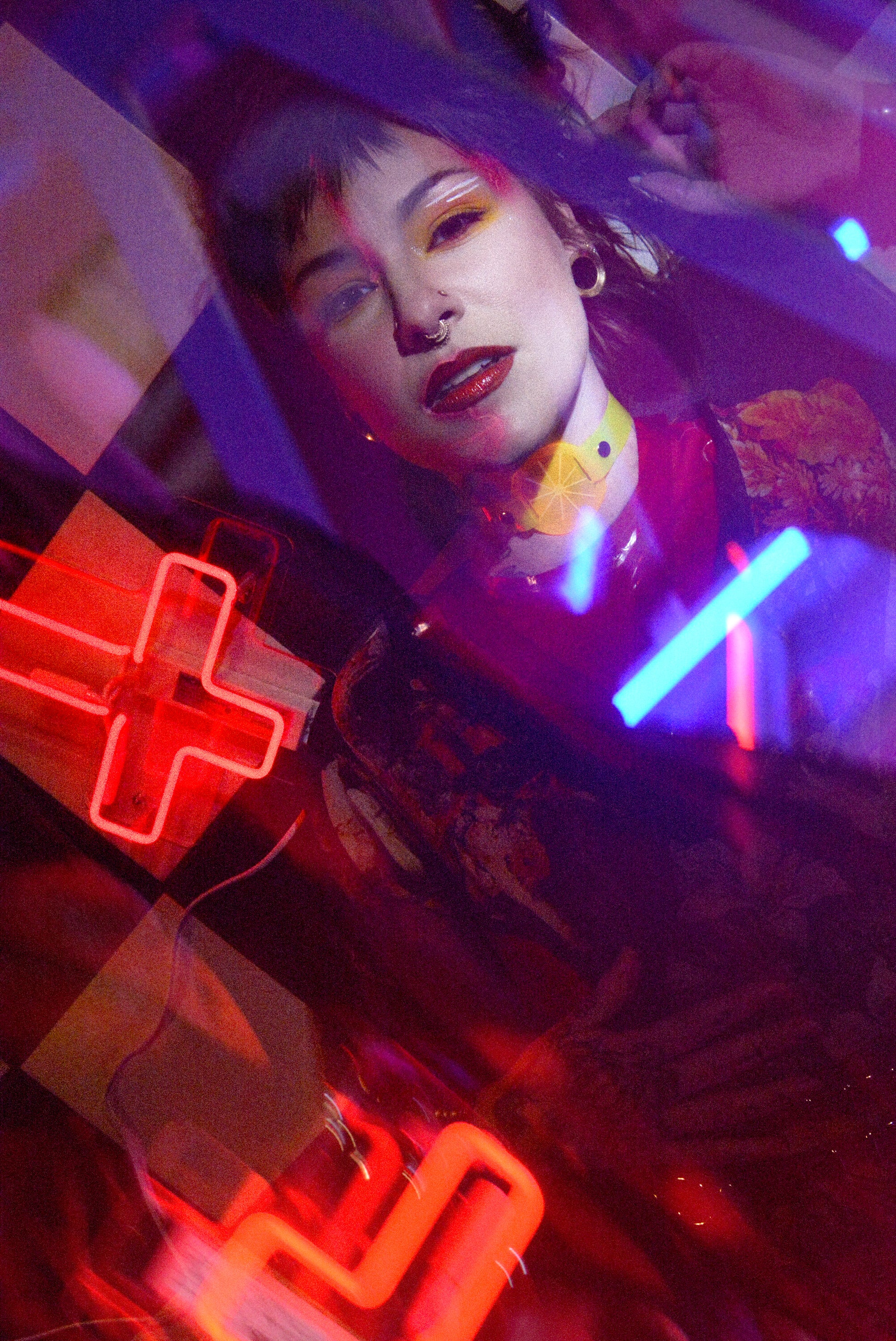 Katrina models a neon uv yellow citrus choker by Apatico and Anhedonie, a floral dress by Samantha Rei, and a red neon cross light under blue and red neon lights at night.