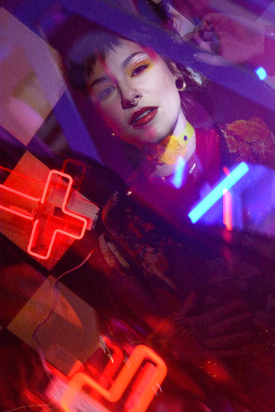 Katrina models a neon uv yellow citrus choker by Apatico and Anhedonie, a floral dress by Samantha Rei, and a red neon cross light under blue and red neon lights at night.