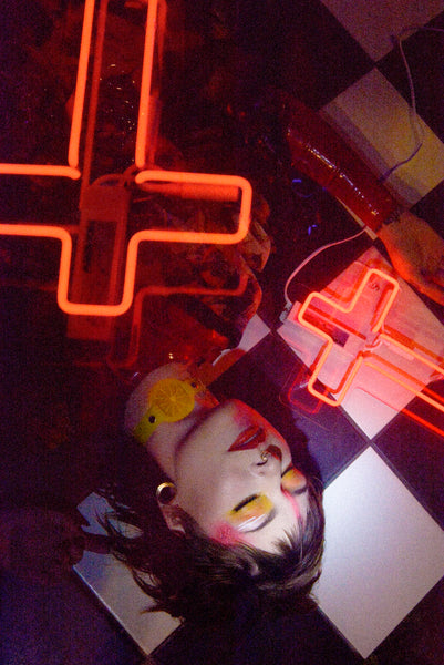  Katrina models a neon uv yellow citrus choker by Apatico and Anhedonie, a floral dress by Samantha Rei, and a red neon cross light under blue and red neon lights at night.