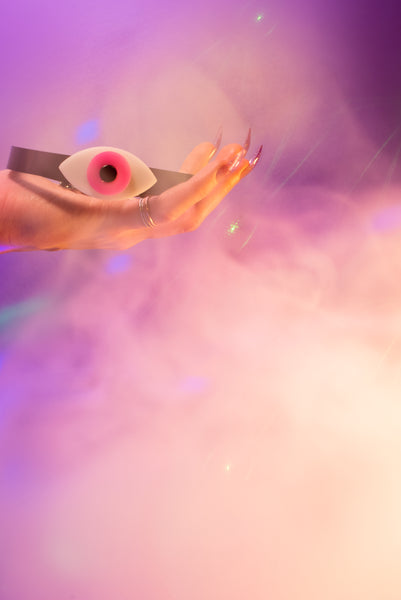 Apatico eyeball choker collar in pink, held in a hand with swirly smoke background.